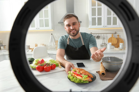 Blogger with tasty croissant sandwich recording video in kitchen at home, view through ring lampの写真素材