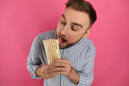 Young man eating delicious shawarma on pink backgroundの写真素材