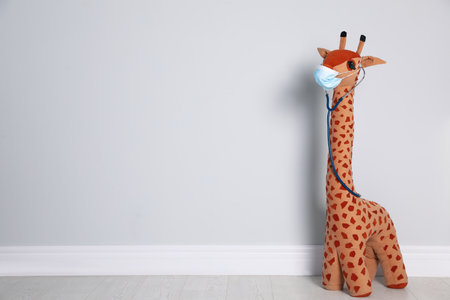 Toy giraffe with face mask and stethoscope near white wall, space for text. Pediatrician practiceの写真素材