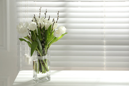 Beautiful bouquet of willow branches and tulips in vase near window, space for textの写真素材