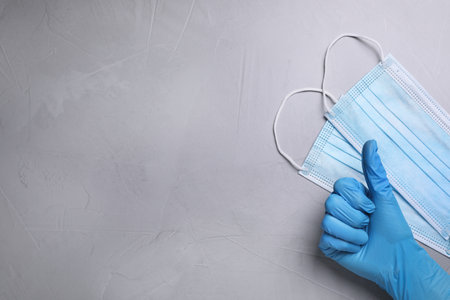 Doctor showing thumb up near protective masks on light gray stone background, top view with space for textの写真素材