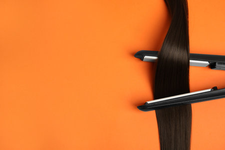 Straightener with brown hair lock on orange background, flat lay. Space for textの写真素材