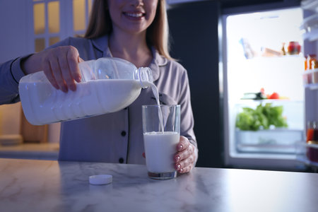Young woman pouring milk from gallon bottle into glass on white marble table in kitchen at night, closeupの写真素材