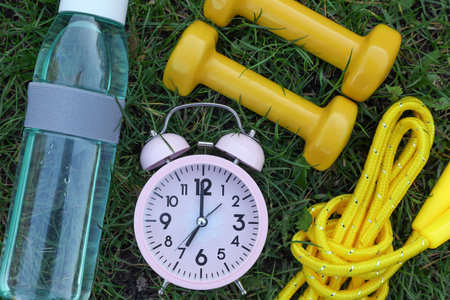 Alarm clock, skipping rope, dumbbells and bottle of water on green grass, flat lay. morning exerciseの写真素材
