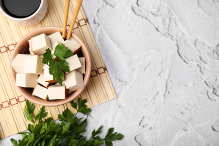 Bamboo mat with bowl of smoked tofu cubes, soy sauce and parsley on white textured table, flat lay. Space for textの写真素材