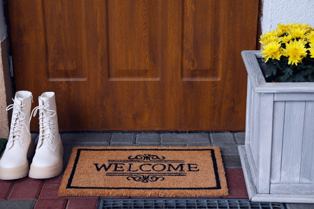 Doormat with word Welcome, stylish boots and beautiful flowers on floor near entranceの写真素材