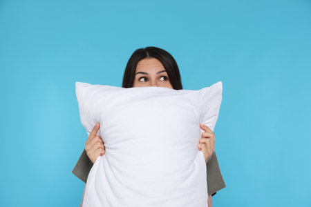 Young woman covering face with soft pillow on light blue backgroundの写真素材