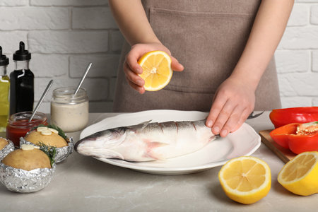 Woman squeezing lemon juice on fresh sea bass fish at light gray marble table, closeupの写真素材