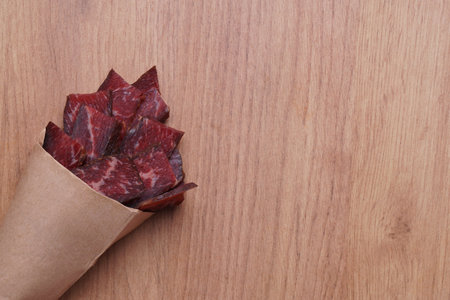 Paper bag with pieces of delicious beef jerky on wooden table, top view. Space for textの写真素材