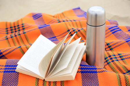 Metallic bottle with hot drink, open book and plaid on sandy beach. closeupの写真素材