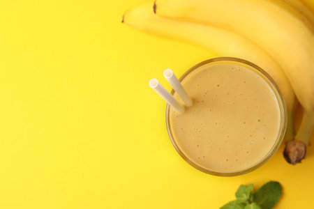 Glass of tasty banana smoothie with straws, fresh fruits and mint on yellow background, flat lay. Space for textの写真素材