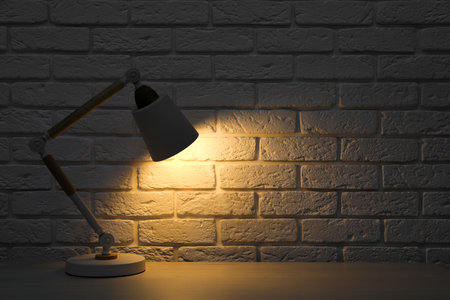Stylish modern desk lamp on white wooden table near brick wall at night, space for textの写真素材