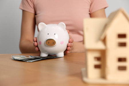 Woman holding piggy bank, house model and banknotes at wooden table, closeupの写真素材