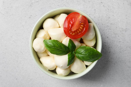 Delicious mozzarella balls in bowl, tomato and basil leaves on light gray tableの写真素材