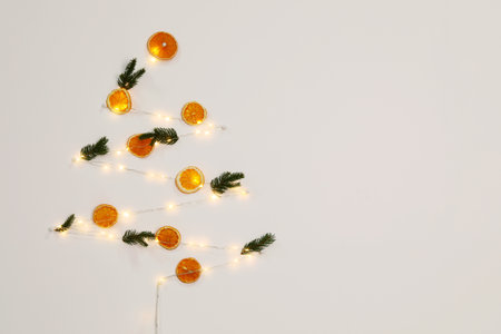 Christmas decor made of dry orange slices, festive lights and fir tree branches on white wall. Space...