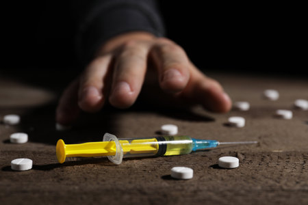 Addicted man reaching to drugs at wooden table, focus on syringeの写真素材