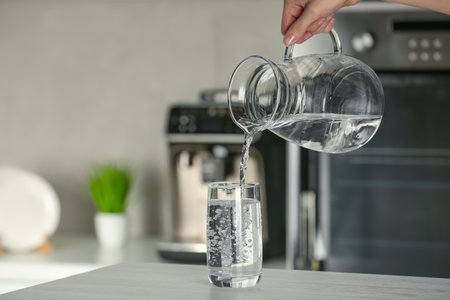 Woman pouring water from jug into glass at wooden table in kitchen, closeup