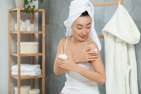 Beautiful young Asian woman applying body cream on shoulder in bathroom, space for textの写真素材