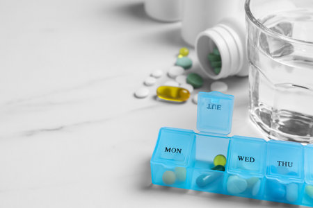 Weekly pill box with medicines and glass of water on white marble table, closeup. Space for textの写真素材