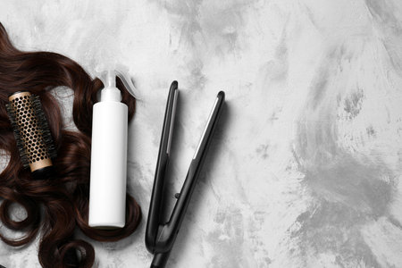 Spray bottle with thermal protection, lock of brown hair, modern straightener and hairbrush on gray...