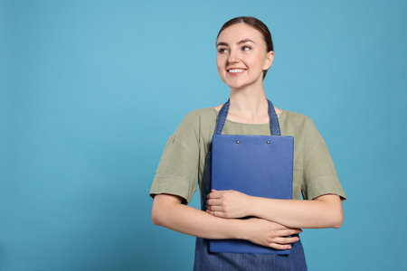 Beautiful young woman in clean denim apron with clipboard on light blue background. Space for textの写真素材