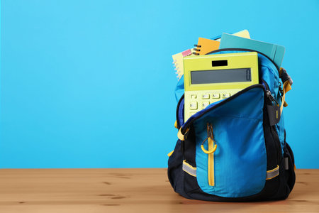 Backpack with different school stationery on wooden table against light blue background, space for...