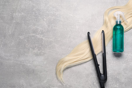 Spray bottle with thermal protection, lock of blonde hair and modern straightener on gray marble table, flat lay. Space for textの写真素材