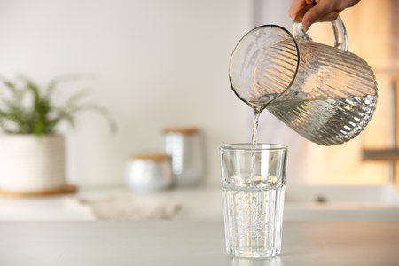 Woman pouring water from jug into glass at white table in kitchen, closeup. Space for textの写真素材