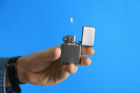 Man holding lighter with burning flame on light blue background, closeup. Space for text
