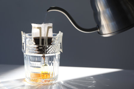 Pouring hot water into glass with drip coffee bag from kettle on light gray table, closeup