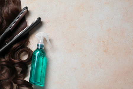 Spray bottle with thermal protection, lock of brown hair and modern straightener on beige textured table, flat lay. Space for textの写真素材
