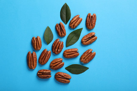 Delicious pecan nuts and green leaves on light blue background, flat lay