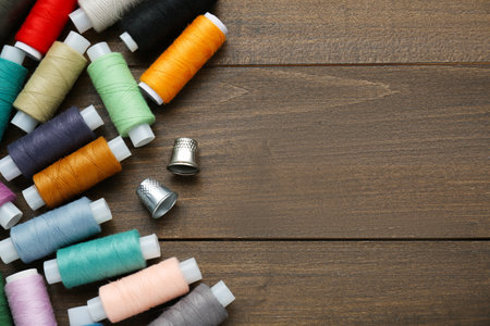 Colorful threads and sewing thimbles on wooden table, flat lay. Space for text