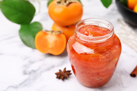 Jar of tasty persimmon jam and ingredients on white marble table. Space for textの写真素材