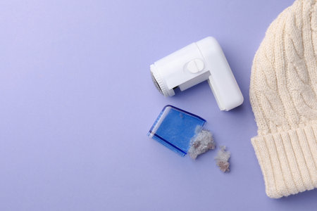 Fabric shaver with fuzz and knitted hat on lilac background, flat lay. Space for textの写真素材