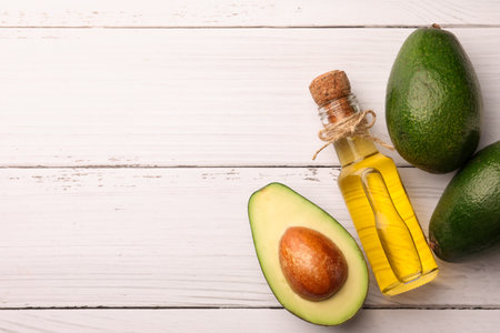 Glass bottle of cooking oil and fresh avocados on white wooden table, flat lay. Space for text