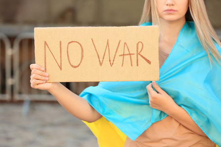 Sad woman wrapped in Ukrainian flag holding poster with words No War against blurred background, closeupの写真素材