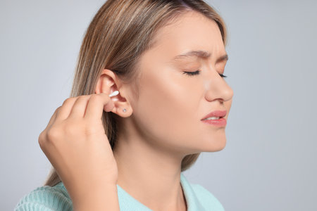 Young woman cleaning ear with cotton swab on light gray background