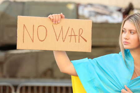 Sad woman wrapped in Ukrainian flag holding poster with words No War outdoorsの写真素材