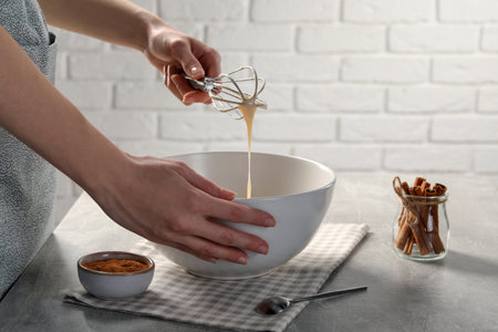 Woman whipping ingredients with whiskey at light gray table, closeup. Cooking delicious eggnog