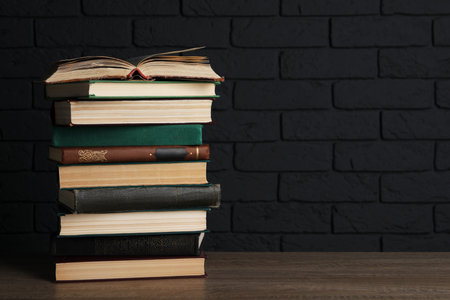 Stack of old hardcover books on wooden table near black brick wall, space for text