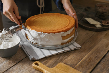 Woman smearing sides of sponge cake with cream on wooden table, closeup