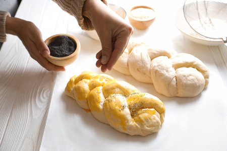 Woman adding poppy seeds onto braided dough at white wooden table in kitchen, closeup. Cooking...