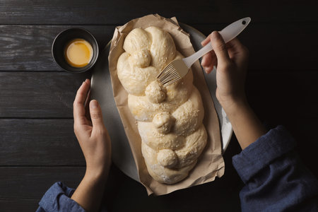 Woman spreading egg yolk onto raw braided bread on black wooden table, top view. Traditional Shabbat...