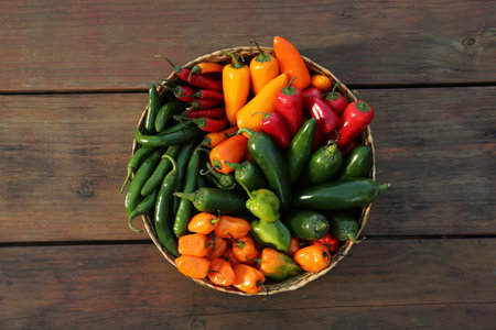 Wicker basket with many different fresh chilli peppers on wooden table, top view