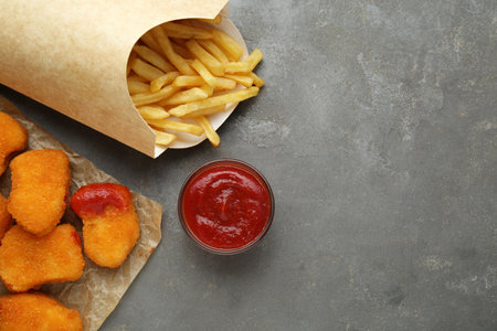 Tasty french fries and chicken nuggets with ketchup on gray textured table, flat lay. Space for text