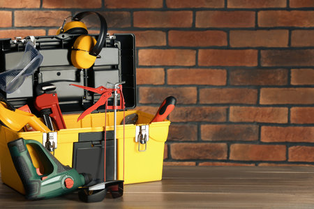 Box with different tools for repair on wooden table near brick wall, space for text