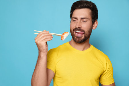 Emotional man holding tasty sushi with chopsticks on light blue backgroundの写真素材