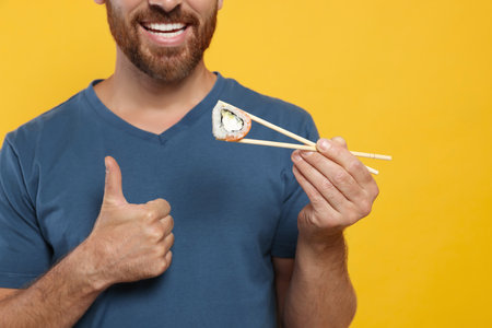 Happy man holding sushi roll with chopsticks and showing thumbs up on orange background, closeupの写真素材