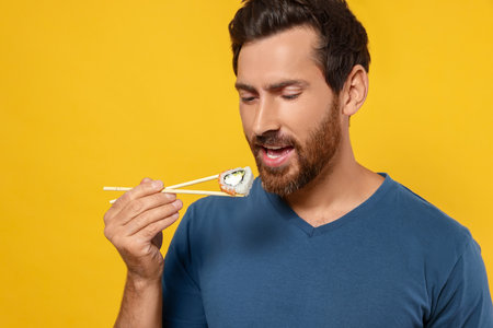 Handsome man eating sushi roll with chopsticks on orange backgroundの写真素材